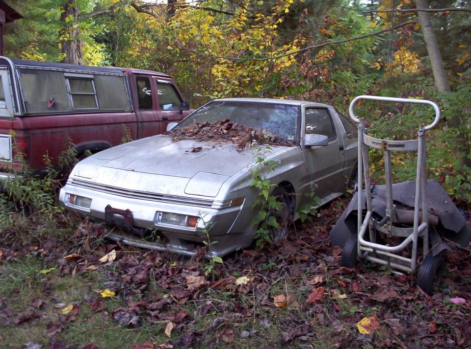 Silver coupe, possibly a Mitsubishi Starion or Chrysler Conquest, overgrown with weeds and leaves on hood. Red pickup truck visible to the left
