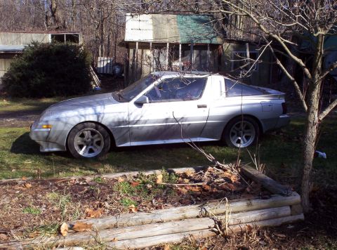 1995 Ford Probe - This silver coupe, resembling a 1995 Ford Probe, is parked outdoors next to a tree and some dilapidated structures.
