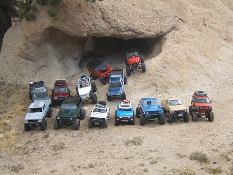 1988 Ford Bronco - Various RC crawlers are gathered on a sandy, rocky landscape, with a large cave-like opening in the background.