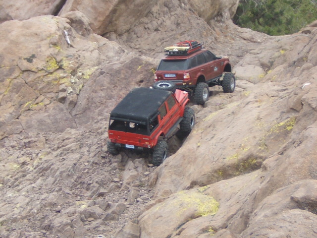 Two remote control off-road vehicles climbing a rocky slope. One is red with a black top, the other is red with a roof rack