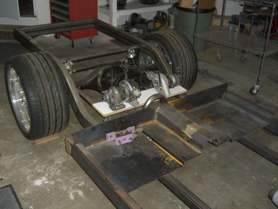 Bare car chassis with rear axle, coilovers, twin turbos and alloy wheels in a garage.