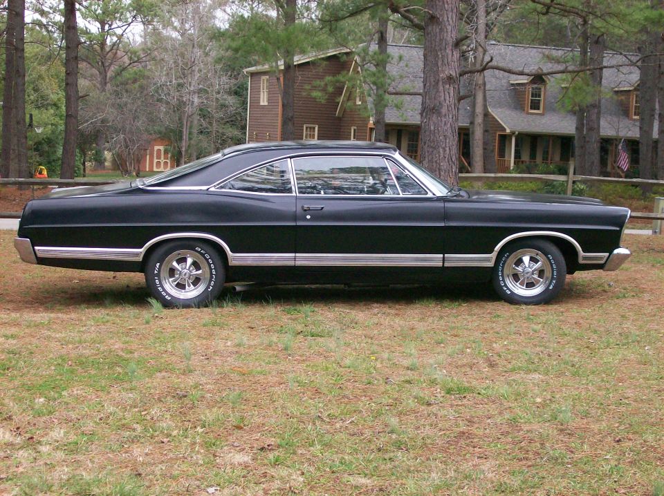 Black Ford Galaxie fastback hardtop side profile on grass with white-letter tires and chrome five-spoke wheels