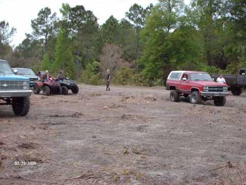 1982 Ford F-100 - Several vehicles, including ATVs and a red SUV, are gathered in a dirt lot with trees in the background.