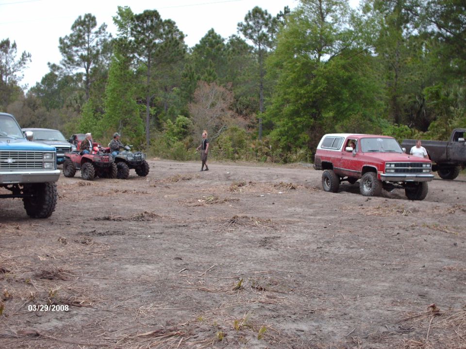 Red SUV with a white top, a blue pickup, and ATVs on a dirt lot