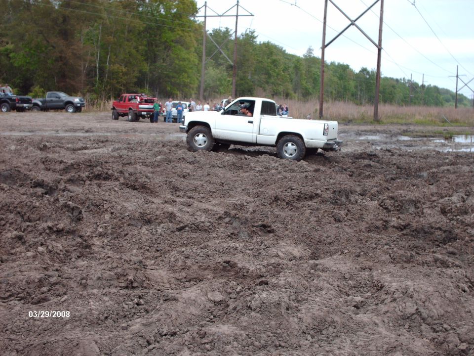 White pickup truck with lifted suspension driving through mud. The truck has a single cab and short bed