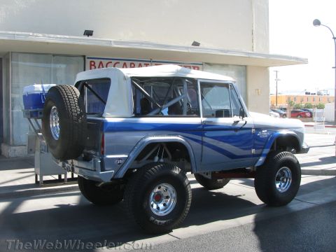 1990 Ford Bronco - lifted blue and white early-model Ford Bronco with a white soft top and rear-mounted spare tire parked by a building.