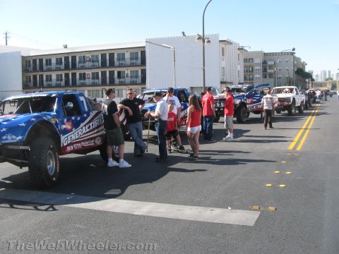 1990 Ford Bronco - line of American-flag-liveried off-road trophy trucks parked along a city street with spectators gathered around.