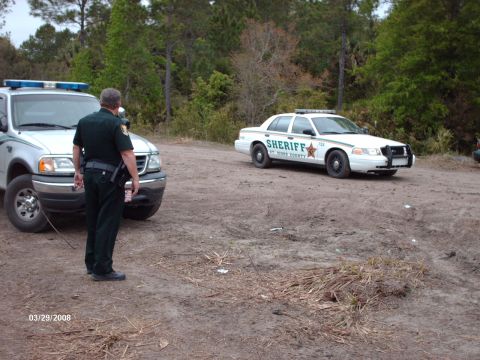 1982 Ford F-100 - Two law enforcement vehicles and an officer are present in a wooded, dirt lot.