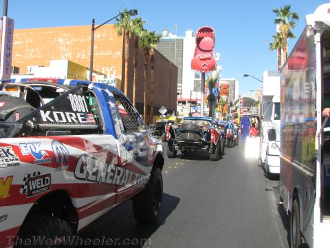 1990 Ford Bronco - Off-road race vehicles including a flag-liveried trophy truck staged along a sunny downtown Las Vegas street.