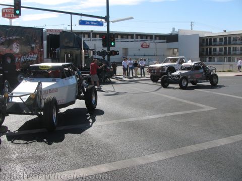 1990 Ford Bronco - Off-road buggies and race trucks staged at a sunny intersection beneath an 8th Street sign with a film camera operator present.
