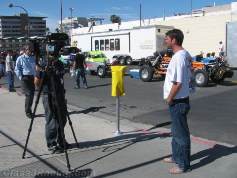 1990 Ford Bronco - cameraman on a tripod films a man being interviewed near off-road race vehicles and an enclosed trailer.