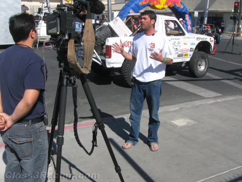 1990 Ford Bronco - man in a white shirt gestures during a filmed interview, with a white off-road race truck and Red Bull arch behind him.