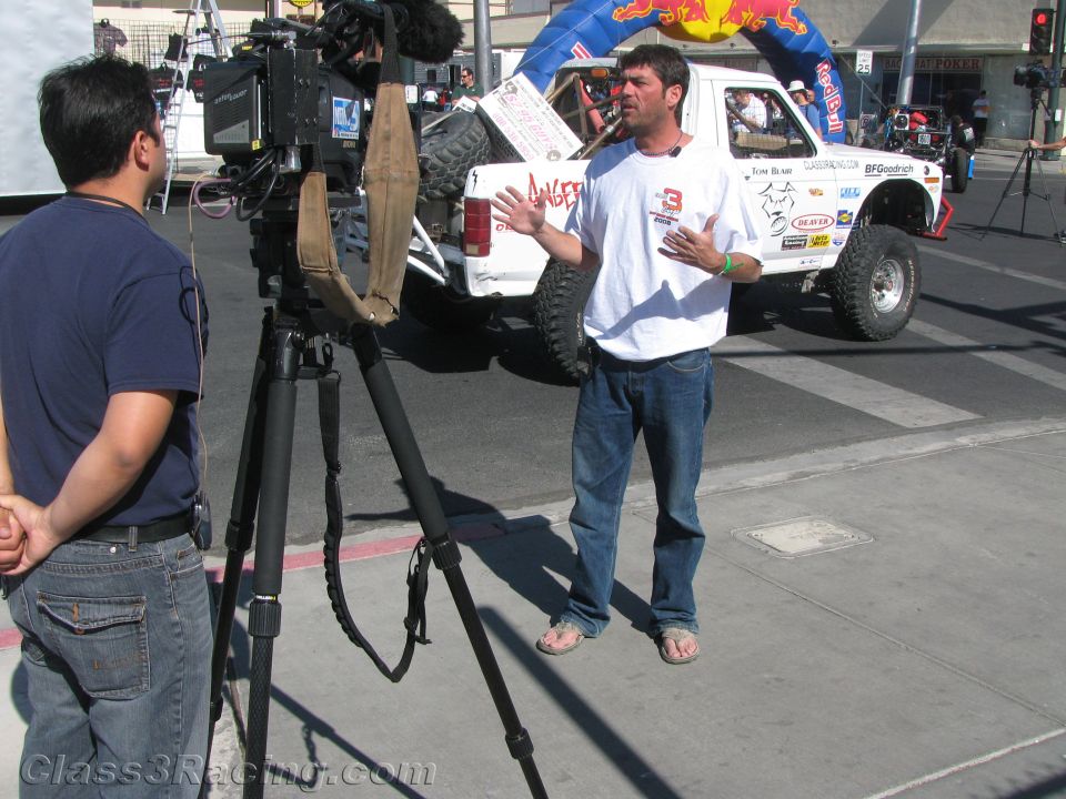 Man gesturing in an interview before a camera with a white race truck behind