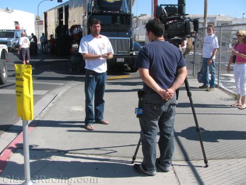 1990 Ford Bronco - man in a white shirt is interviewed by a cameraman on a sunny sidewalk near a parked semi truck and chain-link fence.