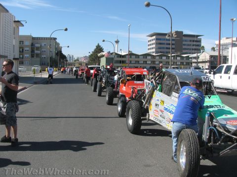 1990 Ford Bronco - long line of off-road race trucks and tube-chassis buggies queued along a sunny city street with spectators alongside.