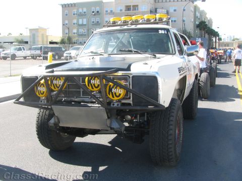 1990 Ford Bronco - white off-road race truck wearing number 361, yellow HID driving lights and a front tube bumper staged on a city street.