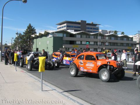 1990 Ford Bronco - orange Baja-style race car numbered 554 lines up with other off-road vehicles on a sunny city street before a multi-story building.