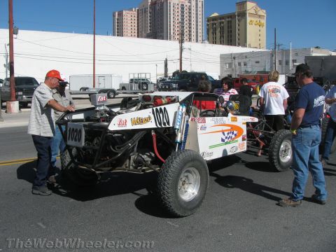 1990 Ford Bronco - Spectators and crew gather around a white tube-frame off-road buggy marked 1020 parked on an urban street with high-rise buildings behind.