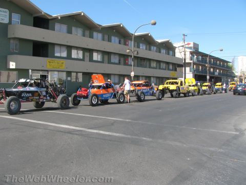 1990 Ford Bronco - long row of colorful off-road buggies and trucks is staged along a street in front of a three-story green apartment building.
