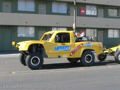 1990 Ford Bronco - yellow tube-frame off-road truck with MESSER and MAXXIS lettering sits on a street in front of a gray building.