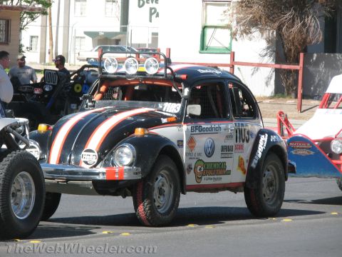 1990 Ford Bronco - Black BFGoodrich-sponsored Baja Bug VW Beetle race car number 1105 with roof light bar at a desert race staging area.