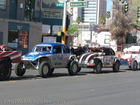 1990 Ford Bronco - blue and a flag-painted orange Baja Bug race car wait at a Seventh Street intersection among other off-road vehicles in a city.