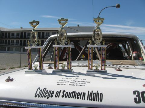 1990 Ford Bronco - Three tall racing trophies stand on the white hood of a College of Southern Idaho race vehicle under a clear blue sky.