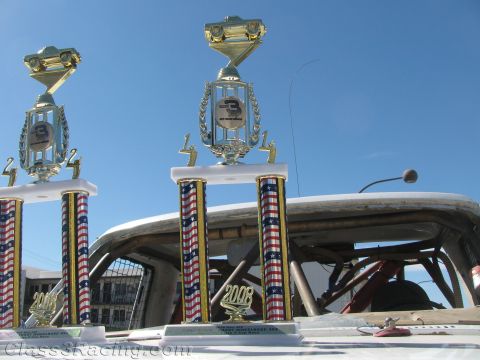 1990 Ford Bronco - Two tall trophies with red-white-and-blue columns and 2008 plates sit atop a white race car against a blue sky.