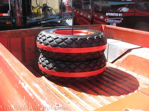 1990 Ford Bronco - Two knobby off-road tires banded with red stripes are stacked upright in the bed of a red truck near a Track Side support rig.