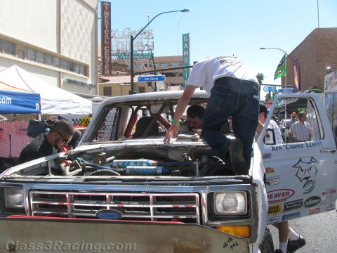 1990 Ford Bronco - Crew members lean over the engine bay of a Ford off-road race truck at a downtown street event with casino signs in the background.