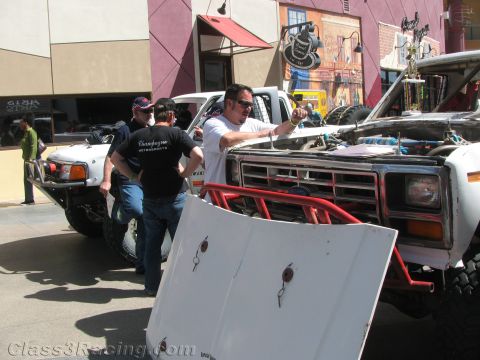 1990 Ford Bronco - Men in t-shirts work around a white off-road race truck with an exposed red roll cage parked on a downtown street.