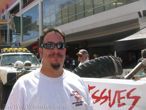 1990 Ford Bronco - man in sunglasses and a gray Class 3 Cup t-shirt poses at a street event with a light-bar race truck and a tire-mounted banner behind.