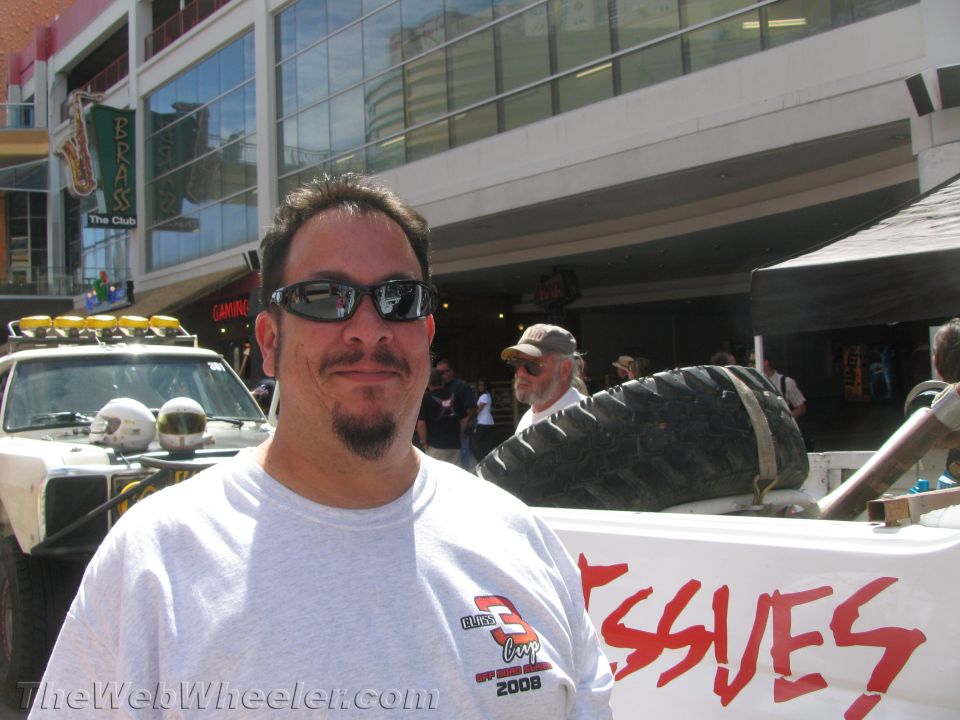Man in sunglasses and gray t-shirt at street event with race truck behind