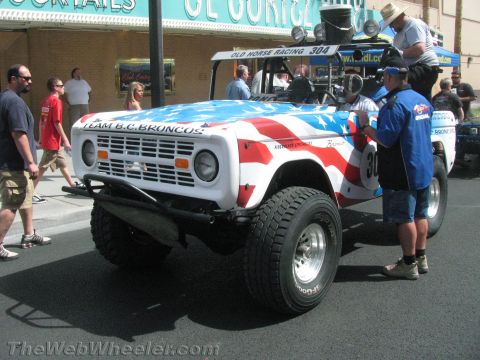 1990 Ford Bronco - white early Ford Bronco race truck numbered 304 with American-flag graphics sits on a street as crew and spectators stand around it.