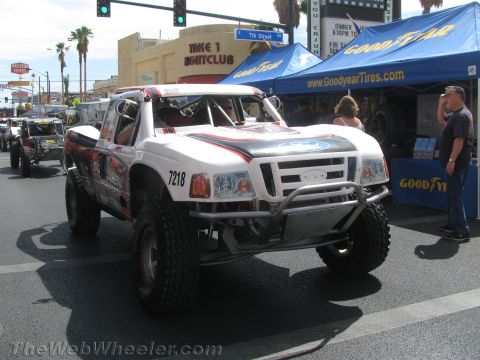 1990 Ford Bronco - white Ford off-road race truck numbered 7218 drives down a street past a blue Goodyear Tires tent and spectators.