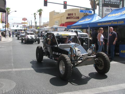 1990 Ford Bronco - black tube-frame off-road race buggy drives down a downtown street past the Take 1 Nightclub marquee and a Goodyear Tires tent.