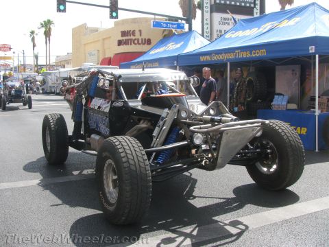 1990 Ford Bronco - black mid-engine tube-frame off-road race buggy with coilover shocks staged on a downtown street near a blue Goodyear Tires tent.