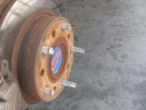 1990 Ford Bronco - Close-up of a rusty vehicle wheel hub and brake rotor with five exposed lug studs and a blue center cap, on a concrete floor.