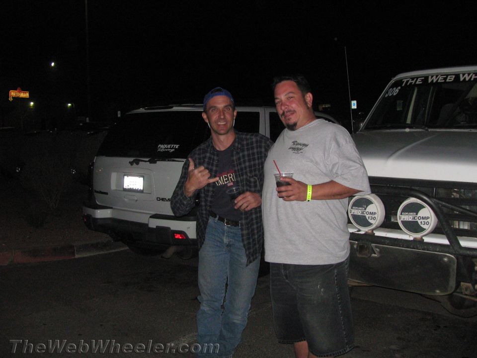 Two men posing at night by white SUVs in a parking lot, one holding a drink