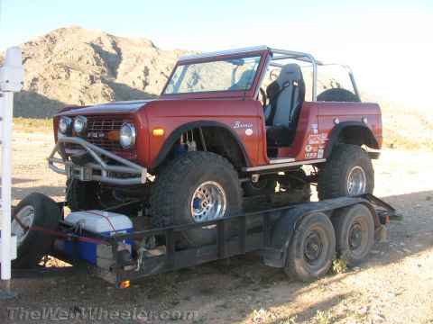 1990 Ford Bronco - lifted red early Ford Bronco with a roll cage and large tires loaded on a tandem-axle trailer in a rocky desert setting.