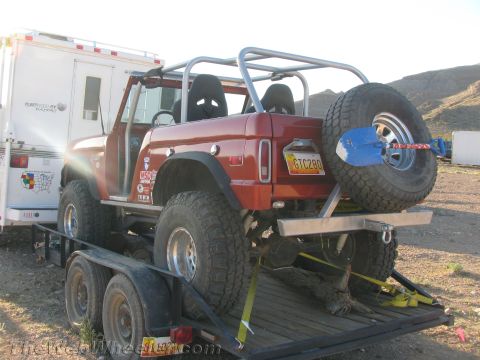 1990 Ford Bronco - Red caged early Ford Bronco off-road buggy with spare tire on rear, strapped to a flatbed trailer beside an RV.