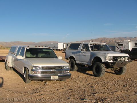 1990 Ford Bronco - white Chevrolet dually pickup and a lifted white Ford Bronco park side by side among trailers and RVs at a dusty desert event.