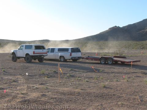 1990 Ford Bronco - Two pickup trucks with camper shells tow an empty flatbed trailer across an open dusty desert flanked by marker stakes.