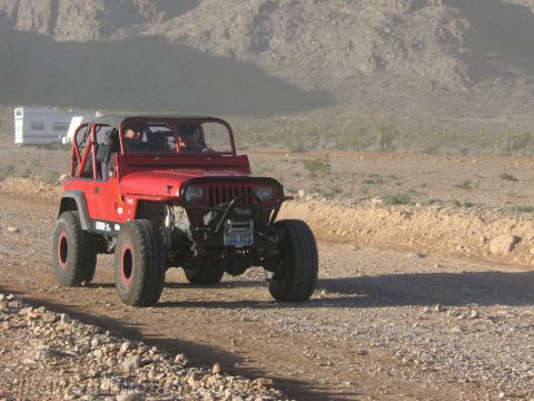 1990 Ford Bronco - red lifted Jeep Wrangler with an open top and roll bar drives along a gravel desert road with a motorhome in the background.