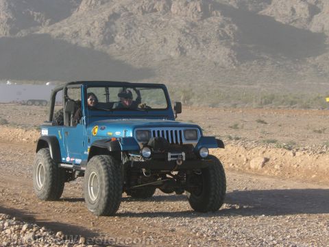 1990 Ford Bronco - blue lifted Jeep Wrangler with an open top, two occupants and a front winch crawls along a gravel trail in mountainous desert terrain.