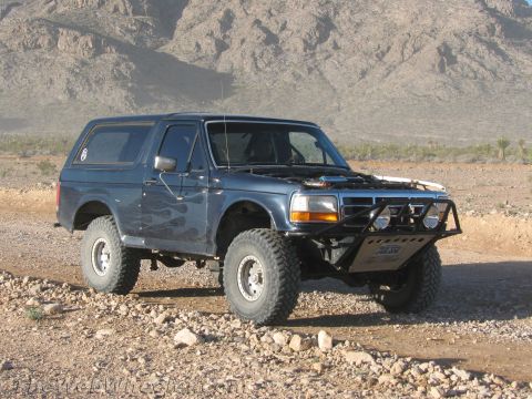 1990 Ford Bronco - dark blue full-size Ford Bronco with camo-style graphics, a tubular front bumper and oversized tires sits on rocky desert ground.