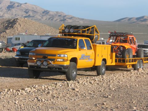 1990 Ford Bronco - yellow Chevrolet crew-cab utility-bed truck with roof-mounted spare tires tows a trailer carrying an orange off-road buggy at a desert even…
