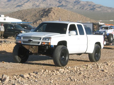 1990 Ford Bronco - white lifted Chevrolet Silverado extended-cab pickup on oversized off-road tires sits among trailers and onlookers at a dusty desert event.