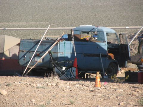 1990 Ford Bronco - old weathered blue cab-over flatbed truck sits at a desert campsite surrounded by tents, poles and an orange traffic cone.