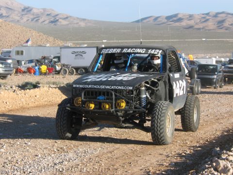 1990 Ford Bronco - black tube-chassis off-road race truck lettered Reber Racing 1421 with yellow auxiliary lights drives a desert course past trailers.